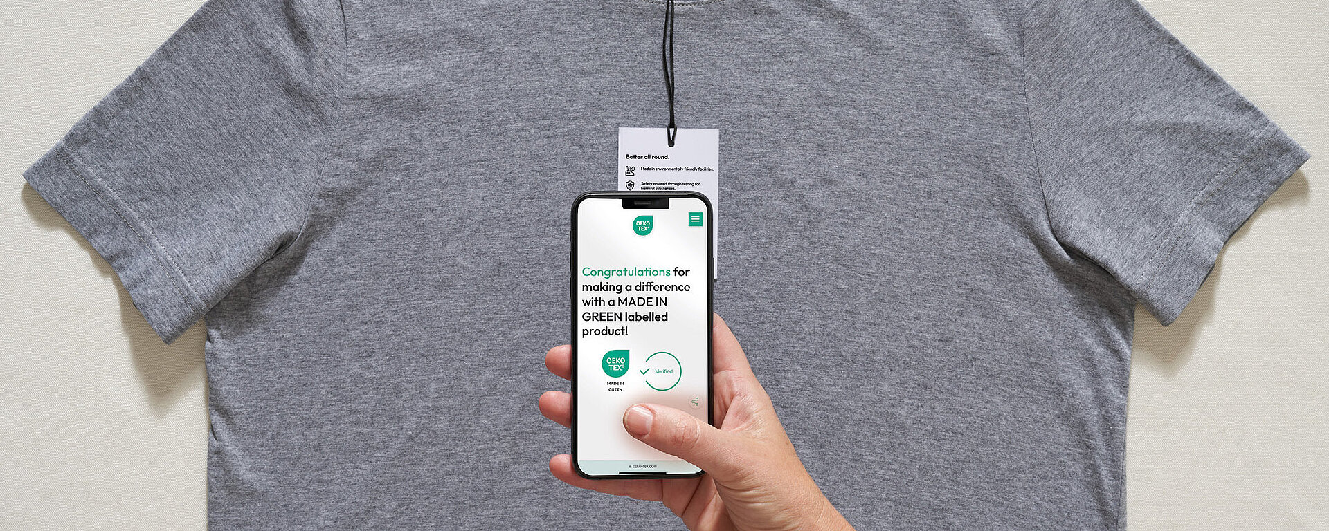 Gray t-shirt with hangtag. Hand holds smartphone showing congratulations message for choosing MADE IN GREEN product.