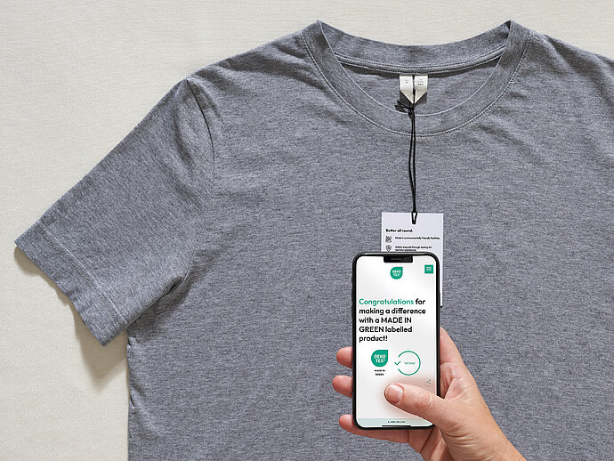 Gray t-shirt with hangtag. Hand holds smartphone showing congratulations message for choosing MADE IN GREEN product.