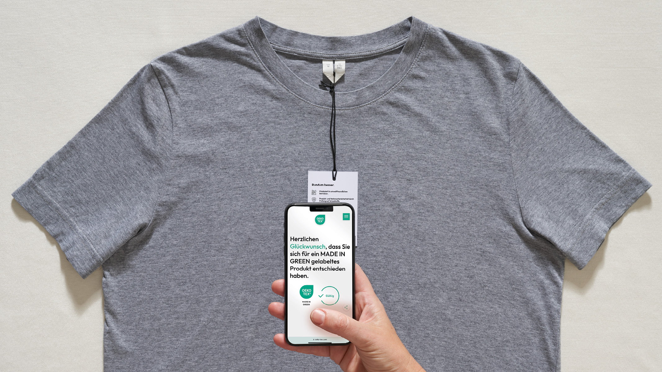 Graues T-Shirt. Hand hält Smartphone mit Glückwunsch-Nachricht für MADE IN GREEN Produktwahl